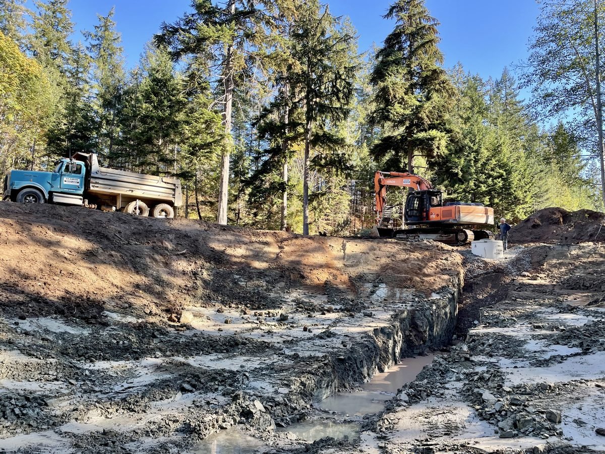 Dump truck and excavator on site during well ring installation, spoil material being placed on surrounding grade